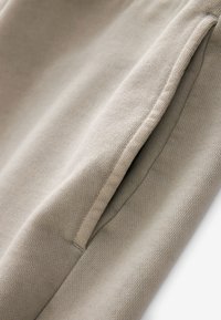 Tessuto beige con una superficie testurizzata, dotato di una tasca laterale visibile e cuciture pulite. Il materiale sembra morbido e flessibile.