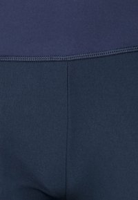 Detailní záběr na tmavě modrou látku s viditelným švem a vyztuženým stehem podél okraje, ukazující texturu a detaily materiálu.