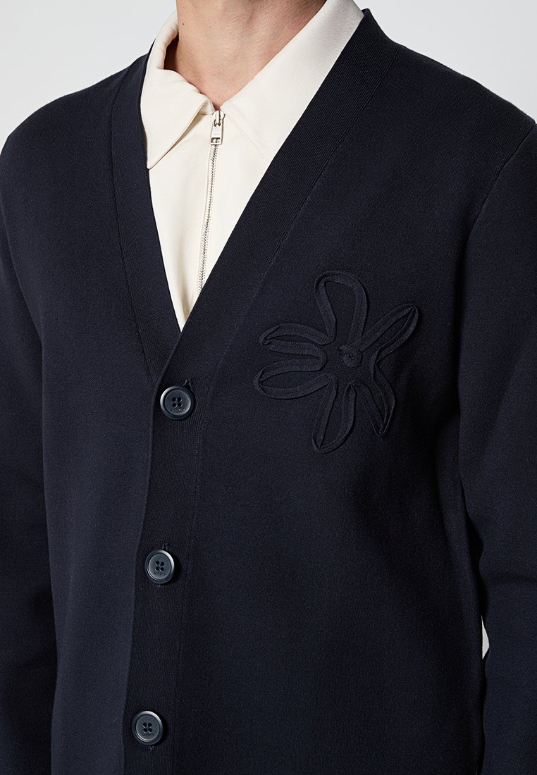 Marineblaue Strickjacke aus weichem Stoff, mit V-Ausschnitt, fünf Knöpfen und einem reliefartigen Blumenmuster auf der linken Brust.