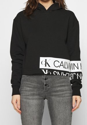 Woman wearing a black Calvin Klein cropped hoodie with white logo bands and grey high-waisted button-fly jeans.