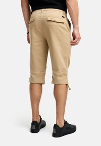 Shorts cargo beiges avec une coupe décontractée, présentant des ourlets repliés, des poches latérales et arrière, et une étiquette près de la taille, associés à des baskets noires.