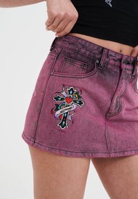 Roze denim minirok met een vervaagde afwerking, voorzien van een opvallend geborduurd kruisdessin met bloemaccenten en een zwarte omranding.