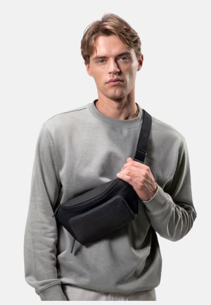 Young man with light brown hair wears gray sweatshirt and black crossbody bag, holding the strap with his right hand.