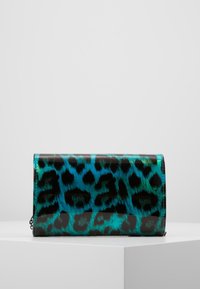 Clutch in pelle con stampa leopardata lucida blu e verde, appoggiata su un piedistallo bianco contro uno sfondo grigio chiaro uniforme.