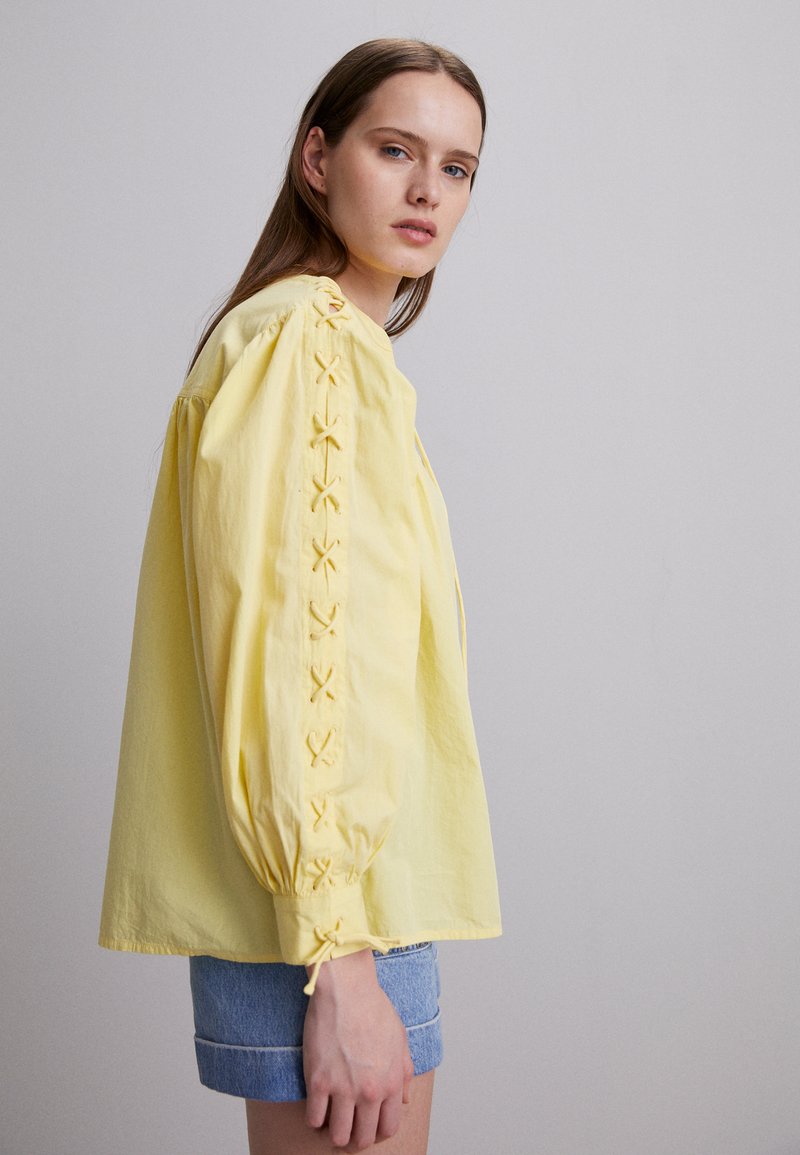 Jeune femme portant un blouse jaune à manches longues avec des détails brodés sur les manches et les poignets, associée à un short en denim retroussé.