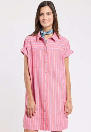 Femme portant une robe à rayures rose et blanche à manches courtes boutonnée et une écharpe bleue nouée autour du cou, souriant à la caméra.