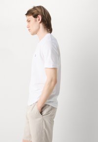 Tricou alb cu maneci scurte, din bumbac, având un mic logo albastru pe piept. Purtați-l cu pantaloni scurți bej deschis.
