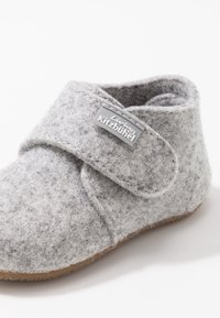 Szare filcowe kapcie z rzepem i beżową gumową podeszwą. Zawierają metkę z logo "Living Kitzbühel". Okrągły kształt, miękka tekstura.