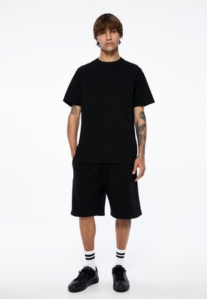 T-shirt en coton noir à manches courtes et col rond, associé à un short noir, porté avec des baskets noires et des chaussettes blanches à rayures noires.