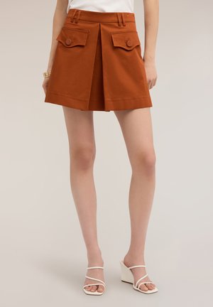 Vrouw draagt oranje geplooide korte broek met knoopzakken en witte sandalen met hak, staand tegen een neutrale achtergrond.