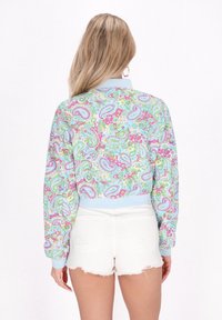 Jachetă bomber cu un model paisley multicolor, cu guler și manșete ribbate albastre, asortată cu pantaloni scurți din denim albi, cu ciucuri.