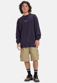 Paars gestreept overhemd met lange mouwen en zwarte manchetten, gecombineerd met beige cargoshorts en zwarte sneakers met oranje accenten.
