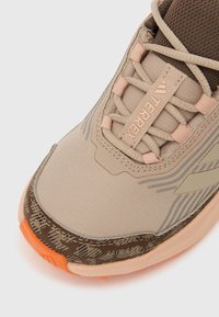 Αθλητικό παπούτσι ανοιχτό μπεζ με υφή στην επιφάνεια, ροζ λεπτομέρειες και καφέ σόλα με σχέδιο. Διαθέτει ευέλικτο σχεδιασμό και σύστημα δέσιμου κορδονιών.