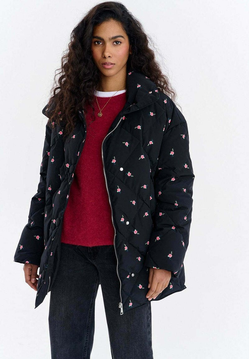 Czarna pikowana kurtka puchowa z wzorem róż, z wysokim kołnierzem, zapinana na zamek błyskawiczny z przodu, z bocznymi kieszeniami, noszona na czerwonym swetrze i czarnych dżinsach.