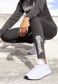 Grijze sweatshirt met "Bench" logo, bijpassende grijze leggings met logo aan de zijkant, en witte sneakers. De outfit is casual en sportief.