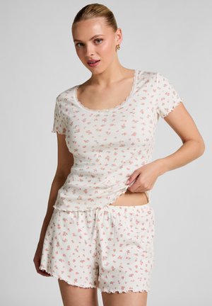 Woman wearing white ribbed short-sleeve top and matching shorts with small pink floral print and lettuce-edge hems, lifting shirt slightly.