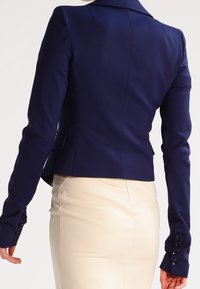 Blazer blu navy aderente con spalle strutturate e maniche lunghe, caratterizzato da bottoni. Abbinato a una gonna beige, che enfatizza il design su misura.