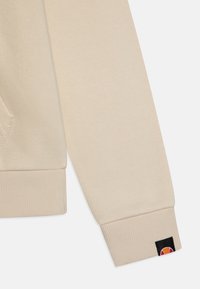 Manga de sudadera beige con puños acanalados, presenta un pequeño parche de logo en negro, naranja y amarillo. Textura suave, diseño casual.