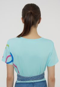 T-shirt azzurro a maniche corte con scollatura rotonda, che presenta forme astratte colorate in rosa, verde e blu sulle spalle.