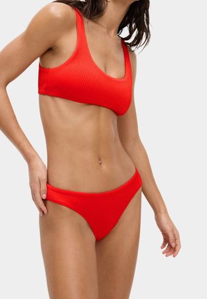 Kobieta w jasnoczerwonym prążkowanym bikini, poprawiająca dolną część stroju, na jednolitym jasnoszarym tle.