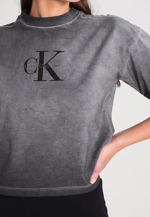 Graues, ausgewaschenes Kurzarmhemd mit schwarzem "Calvin Klein Jeans"-Schriftzug und großem CK-Logo, getragen von einer Person mit Hand in der Hüfte.