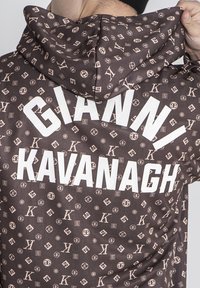 Brun hoodie med ett upprepat logomönster i ljusbeige. Baksidan visar "GIANNI KAVANAGH" i fet stil i vita bokstäver.