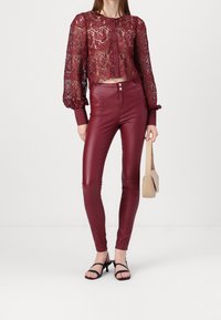 Bordeauxspets topp med ballongsärmar och knäppning, tillsammans med figurnära bordeauxfärgade faux läderbyxor. Modellen håller en beige handväska och bär svarta sandaler.