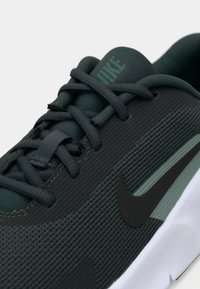 Nærbillede af mørkegrøn Nike løbesko med sort swoosh-logo, tekstureret overflade og sorte snørebånd på en hvid sål.