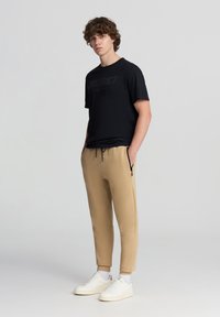 Maglietta nera a maniche corte con la scritta "DISTRICT" in nero, abbinata a jogger beige chiaro a sigaretta con zip e tasche laterali. Sneakers bianche.