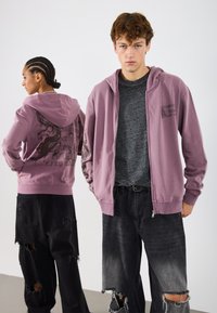 Ροζ φούτερ με φερμουάρ που διαθέτει γραφιστικό σχέδιο στην πλάτη. Συνδυάζεται με μαύρα τζιν με φθορές, παρουσιάζοντας ποικιλία αποχρώσεων και σκισμένα στοιχεία.