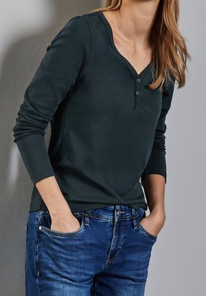 Kvinde iført mørkegrøn langærmet henley-bluse stoppet ned i blå jeans med hænderne i lommerne mod en ensfarvet baggrund.
