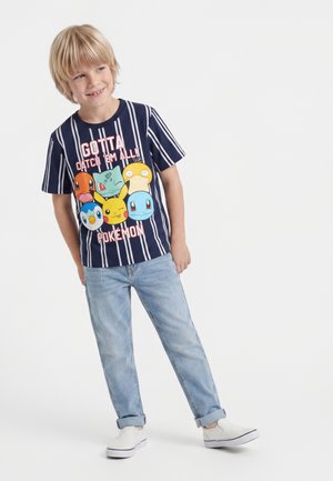 Garçon blond souriant portant un t-shirt Pokémon à rayures bleu marine, un jean bleu clair et des chaussures blanches sans lacets sur fond blanc.