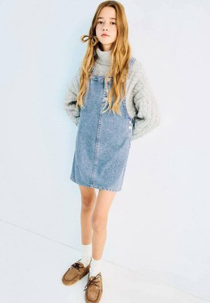 Robe salopette en jean bleu clair avec des boutons en accent, portée avec un pull en maille gris et des mocassins marron. Chaussettes blanches visibles aux chevilles.