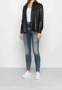 Veste chemise en cuir noir avec un devant boutonné, deux poches poitrine, superposée sur un haut blanc. Assortie avec un jean skinny bleu clair et des baskets blanches.