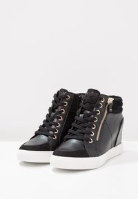 Black high-top sneakers with a mix of leather and suede, featuring gold hardware accents, laces, and a side zipper. White rubber soles.