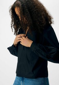 Blusa nera leggera con scollo a V, maniche a sbuffo arricciate e dettagli scallopati ai polsi. Abbinata a jeans di denim.