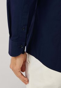 Camisa de manga larga azul marino con puño abotonado y dobladillo curvado, que presenta una textura suave y un diseño de color sólido.