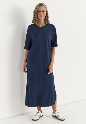 Vrouw met halflang krullend haar, gekleed in een marineblauwe midi-jurk met zijsplit en witte sneakers, staand tegen een effen witte muur.