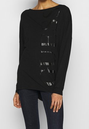 Femme portant un haut noir à manches longues avec un texte vertical brillant et un jean bleu foncé, debout devant un fond uni.
