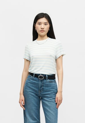 Γυναίκα φοράει ανοιχτό μπλε και άσπρο ριγέ κοντομάνικο πουκάμισο φορεμένο μέσα σε ψηλόμεσα μπλε τζιν με μαύρη ζώνη σε λευκό φόντο.