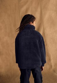 Veste en polaire bleu marine avec col haut, tissu texturé et épaules basses. Détail de patch carré notable dans le dos.