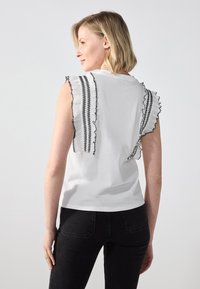 White sleeveless top with black ruffled detailing along shoulders, featuring zigzag stitching; worn with dark jeans in a plain background.
