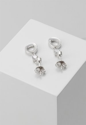 Boucles d'oreilles clous en argent avec anneaux ovales, petites pierres précieuses transparentes et fermoirs papillon, exposées sur un socle cubique blanc.