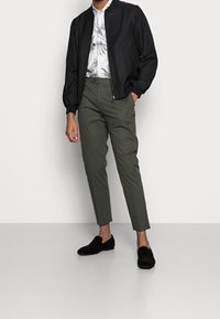 Černá bomber bunda, bílá květinová košile, olivově zelené kalhoty a černé semišové mokasíny. Minimalistický design s dobře padnoucím střihem a hladkou texturou.