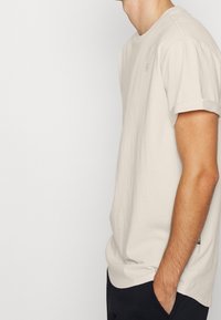 Homme portant un t-shirt beige clair et un pantalon noir, debout de profil avec une main dans la poche sur un fond blanc uni.