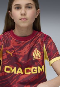 Röd och gul fotbollströja med ett vågmönster, med Puma-logga och Marseille-emblem. Texten lyder "CMA CGM" i fet gul.