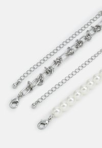 Trois chaînes : une en argent avec des maillons entrelacés, une délicate chaîne en argent, et un fil de perles blanches, toutes avec des fermoirs à homard.