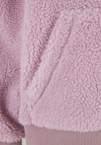Roze polar jakna s teksturiranim, pahuljastim završetkom. Ima rebraste manžetne u svjetlijoj roze boji, koje naglašavaju meku, udobnu tkaninu.