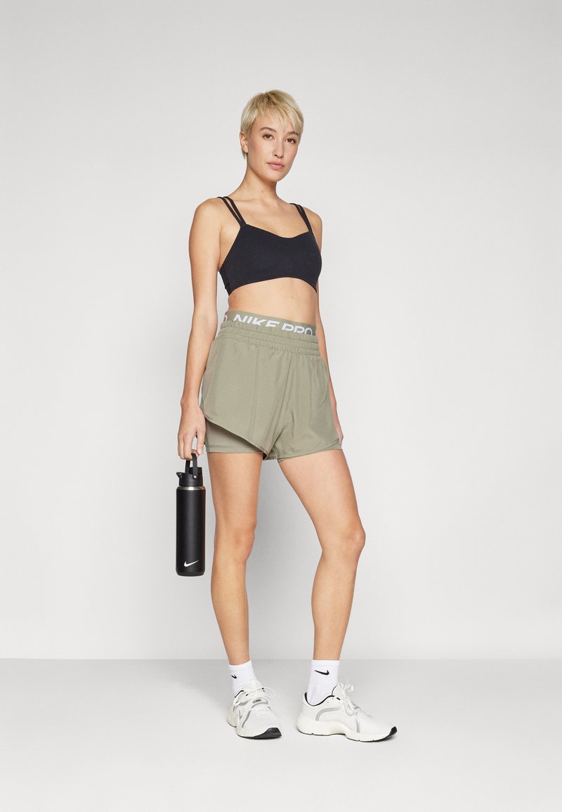 Top corto atlético negro, pantalones cortos verde oliva con cintura "NIKE PRO", zapatillas blancas, botella de agua negra en la mano, fondo liso.