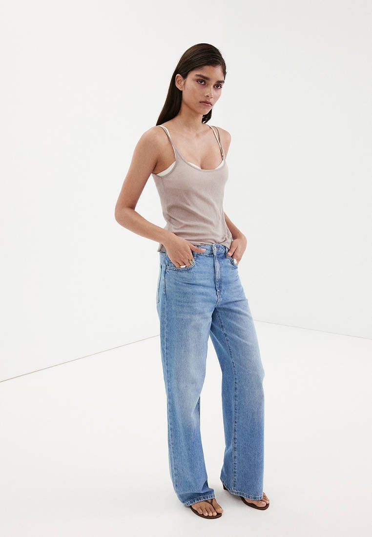 Femme debout, les mains dans les poches, portant un top beige sans manches, un jean large bleu clair et des sandales foncées sur un fond blanc uni.